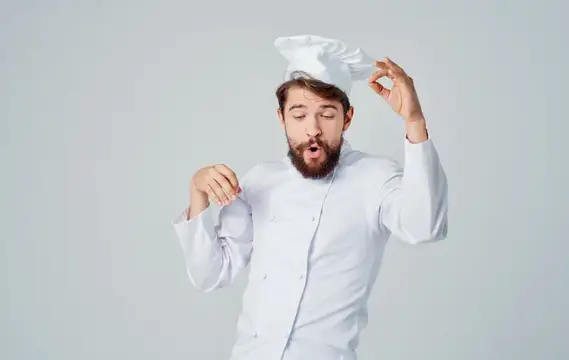 Бородатый повар в белом колпаке жестикулирует на сером фоне