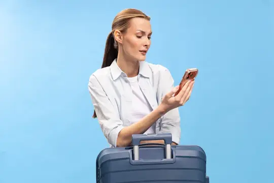 Светловолосая женщина с чемоданом смотрит в смартфон на голубом фоне