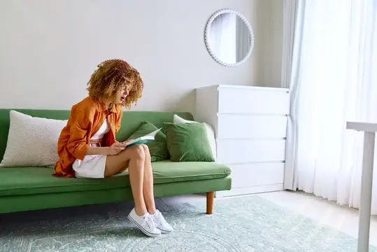 Кудрявая девушка в оранжевой рубашке читает книгу на зелёном диване