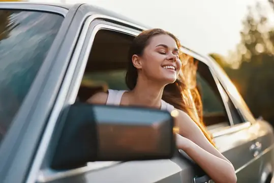 Молодая женщина с закрытыми глазами высунулась из окна автомобиля на закате