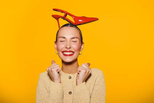 Улыбающаяся девушка с красной туфлей на голове на жёлтом фоне