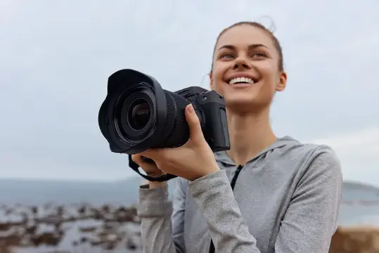 Счастливая молодая женщина с фотоаппаратом на пляже, одетая в повседневную спортивную одежду, наслаждается временем на свежем воздухе