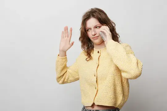 Молодая женщина в желтом свитере прижимает смартфон к уху и машет рукой в знак приветствия