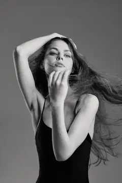 Чёрно-белый портрет молодой женщины с длинными развевающимися волосами и безмятежным выражением лица, одетой в простой чёрный топ на однотонном фоне