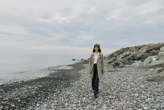 Молодая женщина в пальто и кепке стоит на каменистом берегу моря
