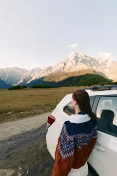 Женщина опирается на припаркованный автомобиль и смотрит на горные вершины и открытый пейзаж