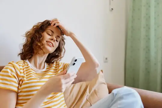 Кудрявая девушка в полосатой футболке отдыхает со смартфоном дома