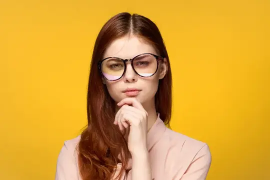 Рыжеволосая девушка в очках задумчиво смотрит на жёлтом фоне