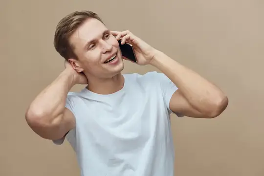 Улыбающийся мужчина в повседневной белой футболке держит смартфон у уха, разговаривая по телефону в расслабленной позе на бежевом фоне, концепция цифрового кочевника, иллюстрирующая образ жизни…