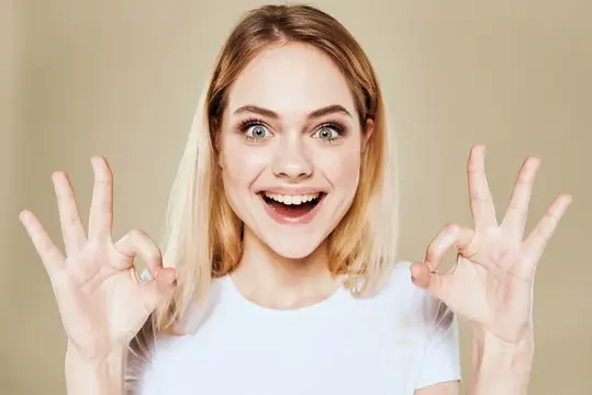 Весёлая блондинка в белой футболке жестикулирует рукой, выражая эмоции на бежевом фоне