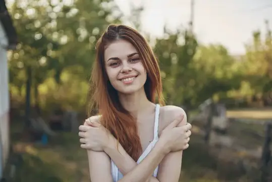 Рыжеволосая девушка улыбается и обнимает себя на летнем дворе