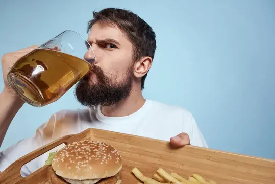 Бородатый мужчина пьёт пиво с бургером и картошкой