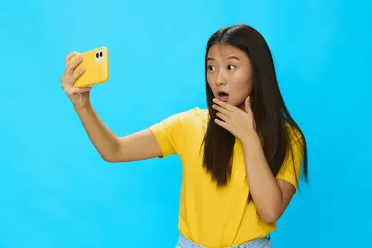 Удивлённая азиатская девушка делает селфи на жёлтый смартфон на голубом фоне
