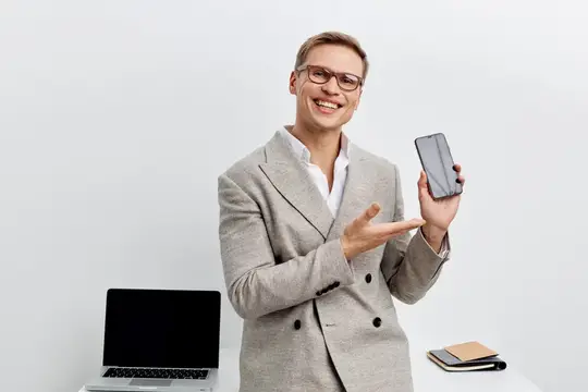 Улыбающийся мужчина в очках показывает смартфон на фоне ноутбука