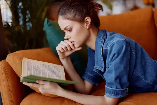 Женщина отдыхает и читает книгу, устроившись на удобном диване в уютной гостиной; на заднем плане — зелёное растение