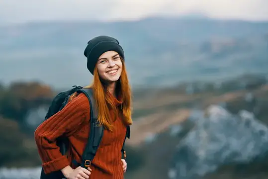 Улыбающаяся рыжеволосая девушка с рюкзаком на фоне гор