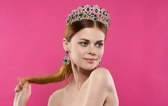 Молодая женщина в роскошной короне и серьгах на розовом фоне