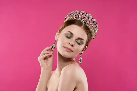 Молодая женщина в роскошной короне и серьгах на розовом фоне