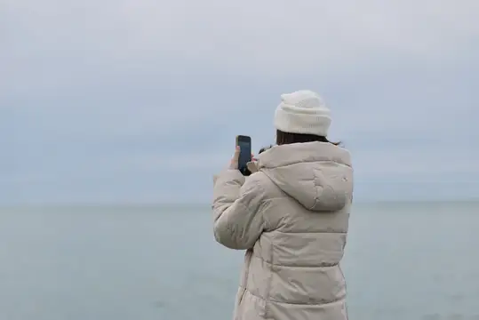 Женщина в светлом пуховике фотографирует море на телефон