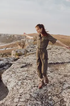 Приключение на природе: женщина в зелёном комбинезоне триумфально стоит на скалистом выступе, раскинув руки в стороны
