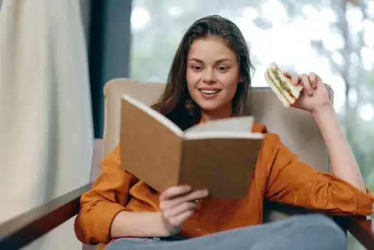Счастливая молодая женщина в оранжевой рубашке читает книгу, одновременно наслаждаясь бутербродом
