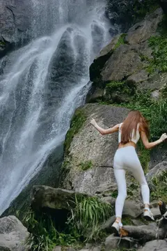 Девушка в белом стоит на камнях у подножия водопада