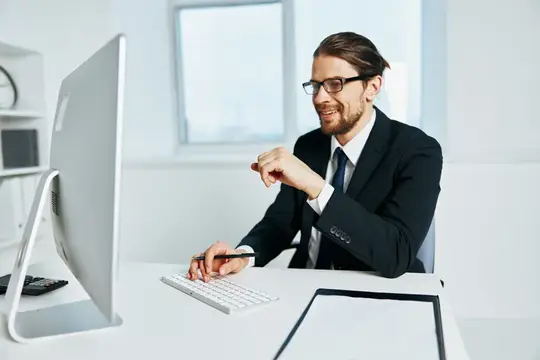 Улыбающийся бородатый мужчина в очках и костюме работает за компьютером в офисе