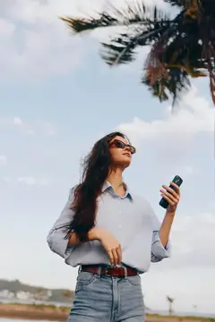 Девушка в солнцезащитных очках с телефоном на фоне пальмы и неба