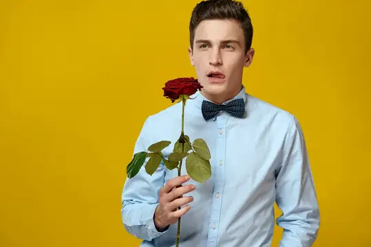 Молодой человек в голубой рубашке с бабочкой держит красную розу на жёлтом фоне