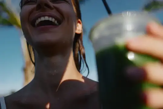 Смеющаяся девушка с зеленым смузи в руке на фоне пальм