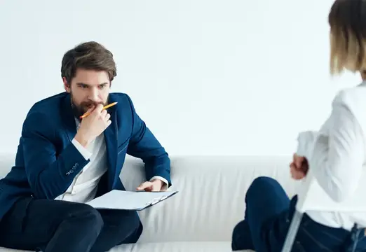Бородатый мужчина в синем пиджаке слушает клиентку на белом диване