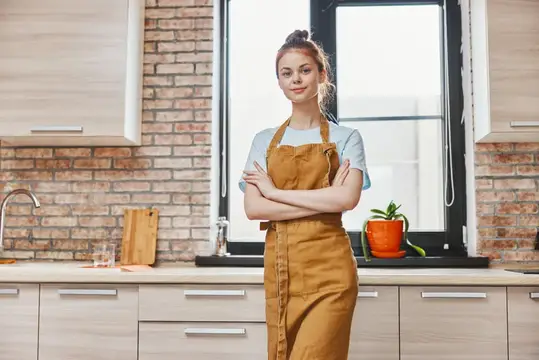 Молодая женщина в фартуке стоит со скрещёнными руками на кухне