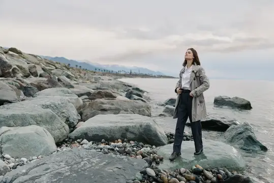 Молодая женщина в плаще стоит на камнях у моря в пасмурный день