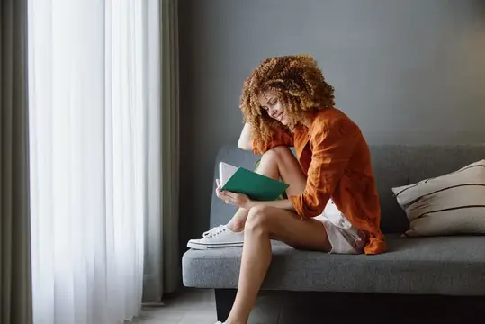Кудрявая девушка в оранжевой рубашке читает книгу на диване у окна