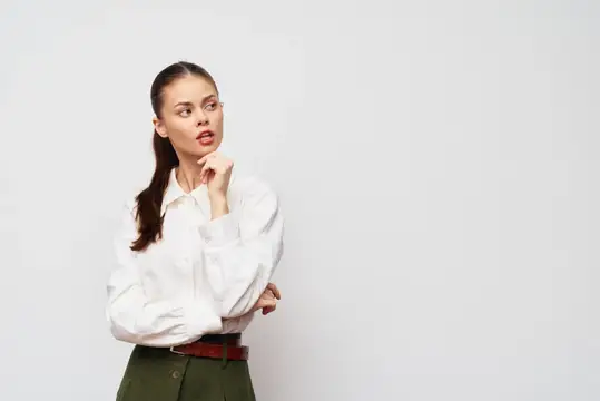 Задумчивая женщина, приложившая руку к подбородку, одетая в белую блузку и оливково-зелёные брюки, стоит на нейтральном светло-сером фоне, передавая ощущение размышления и любопытства о будущем