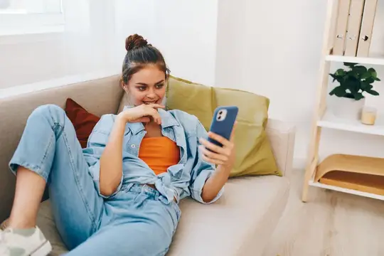 Улыбающаяся девушка лежит на диване со смартфоном в руках