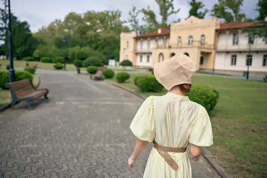 Женщина в соломенной шляпе и кремовом платье идёт по аллее у старинной усадьбы