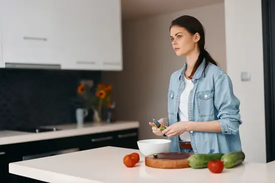 Молодая женщина в джинсовой куртке режет овощи на светлой кухне