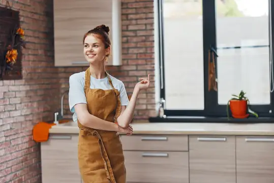 Улыбающаяся рыжеволосая девушка в фартуке стоит на кухне у окна