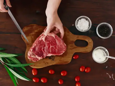 Руки нарезают сырое мясо на деревянной доске с помидорами и специями