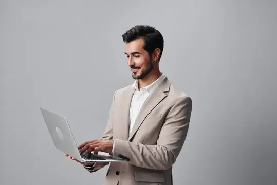 Улыбающийся мужчина в бежевом пиджаке работает на ноутбуке на сером фоне