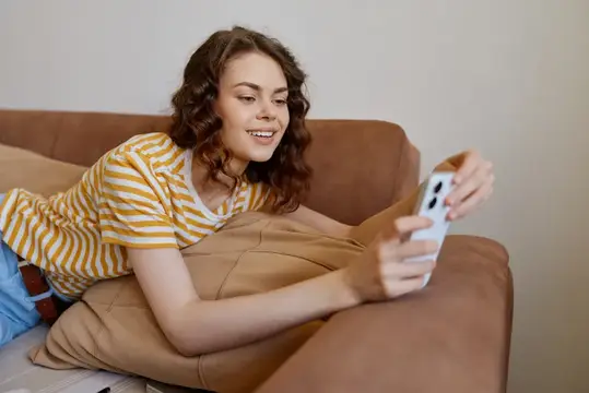 Кудрявая девушка лежит на диване и улыбается, глядя в смартфон