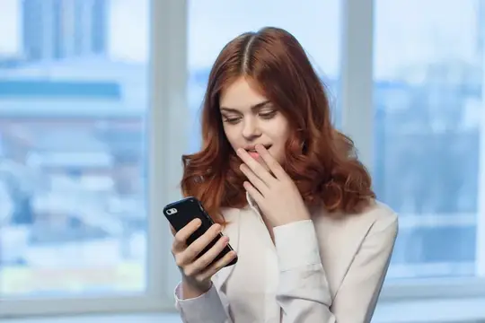 Рыжеволосая девушка со смартфоном удивлённо прикрывает рот рукой у окна