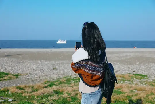 Темноволосая девушка фотографирует море и корабль на телефон