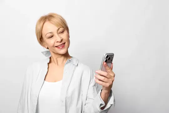 Улыбающаяся блондинка в белой рубашке смотрит в смартфон на светлом фоне