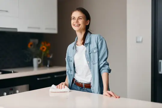 Улыбающаяся девушка в джинсовой рубашке вытирает стол на светлой кухне
