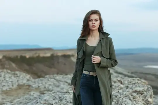 Молодая женщина в оливковом плаще стоит на каменистом холме