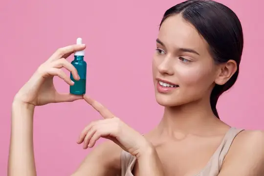 Улыбающаяся темноволосая девушка держит флакон сыворотки на розовом фоне