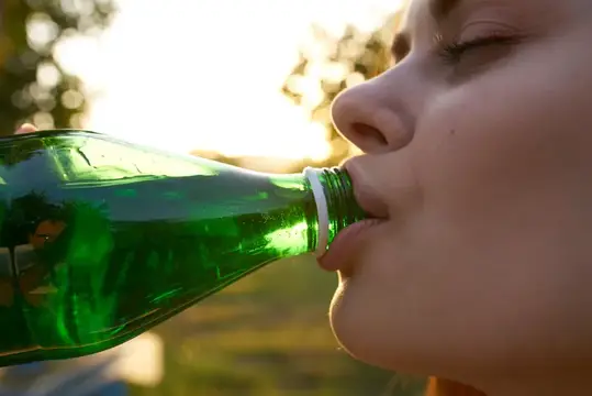 Девушка пьёт воду из зелёной стеклянной бутылки на закате