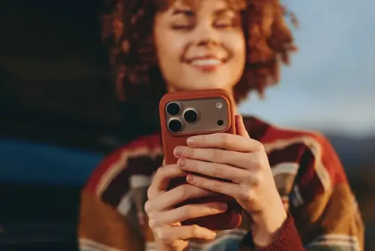 Улыбающаяся рыжеволосая девушка держит смартфон в красном чехле